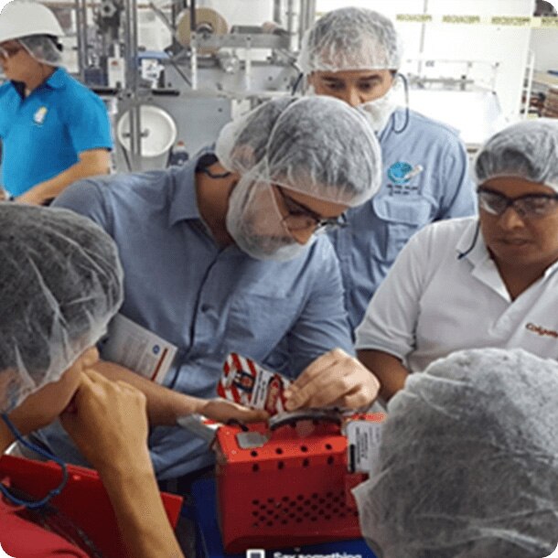 Un grupo de trabajadores con cofias de protección e indumentaria industrial observan atentamente a un colega que manipula un componente o equipo en una planta de producción.