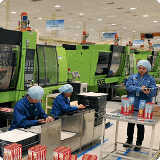Tres operarias con uniformes azules y cofias protectoras trabajan en una línea de producción industrial con grandes maquinarias verdes, empacando y revisando productos de higiene bucal.